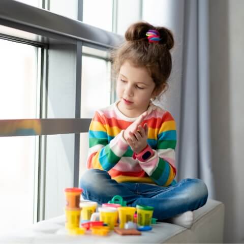 5-year-old girl sitting on a window frame and playing with toys.