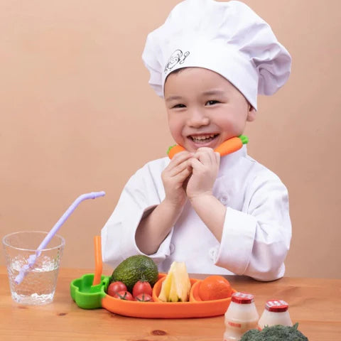 Little boy in a kitchen chef outfit eating fruits and vegetables from "My First Dining Set" made out of silicone. 