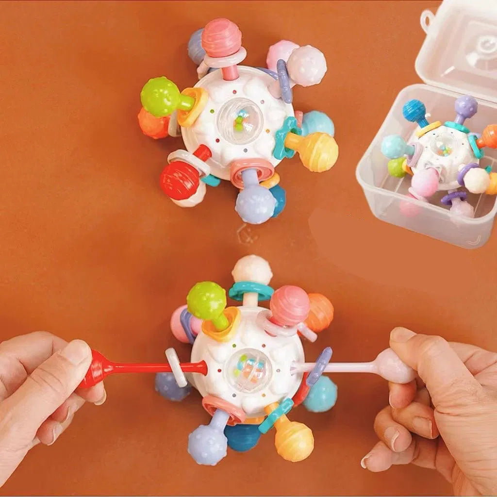 A colorful sensory teething toy with multiple textured handles being pulled apart by two hands. The toy features various soft, rounded extensions and a central rattle ball, designed for tactile exploration and teething relief. 